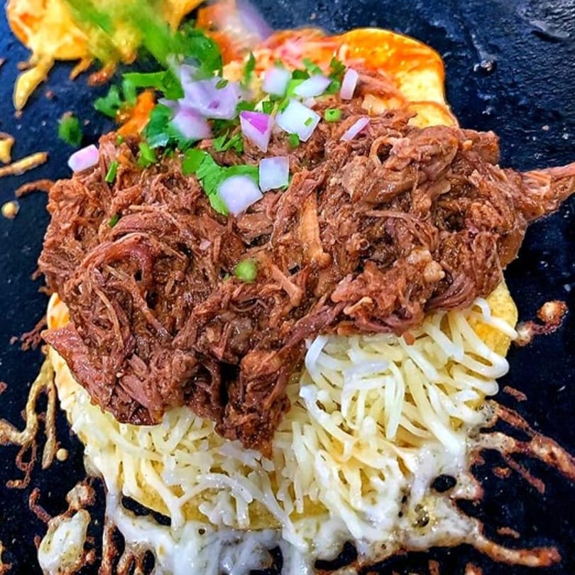 Close-up of a birria taco with shredded beef, melted cheese, onions, and cilantro on a griddle.