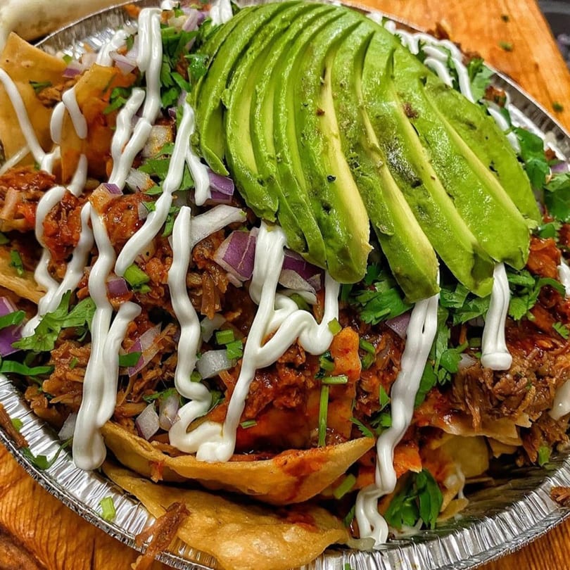 La Baja birria nachos with sliced avocado, sour cream, red onion, and cilantro in a foil container.